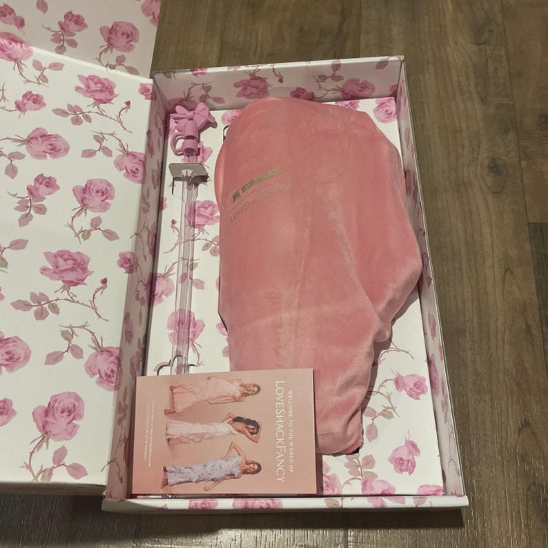 Op de afbeelding: Open doos met een roze rozenpatroon. Binnenin zijn een roze fluwelen kledingzak en een doorzichtige glazen kledinghanger met een roze strik zichtbaar. Een kleine kaart met de tekst "LOVESHACKFANCY" staat ook in de doos.