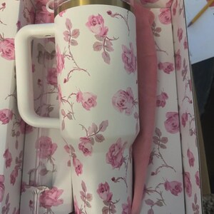 Op de afbeelding: Een witte ge&iuml;soleerde beker met een handvat, versierd met roze rozenpatronen. De beker heeft een gouden rand en een doorzichtig deksel. Het wordt gepresenteerd in een doos met een bijpassend bloemmotief, vergezeld van een roze hoes en een doorzichtig rietje.