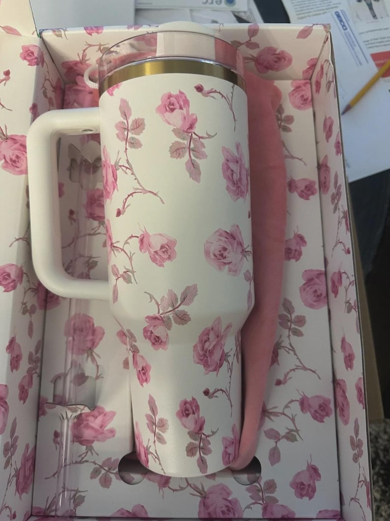 Op de afbeelding: Een witte ge&iuml;soleerde beker met een handvat, versierd met roze rozenpatronen. De beker heeft een gouden rand en een doorzichtig deksel. Het wordt gepresenteerd in een doos met een bijpassend bloemmotief, vergezeld van een roze hoes en een doorzichtig rietje.