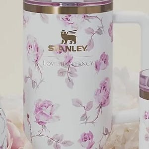 Op de afbeelding: Witte Stanley-beker met een bloemenrozenpatroon in roze en grijstinten. De beker heeft een handvat, een doorzichtig deksel en een decoratieve strik op het rietje. De logo's van Stanley en Love Shack Fancy zijn zichtbaar.