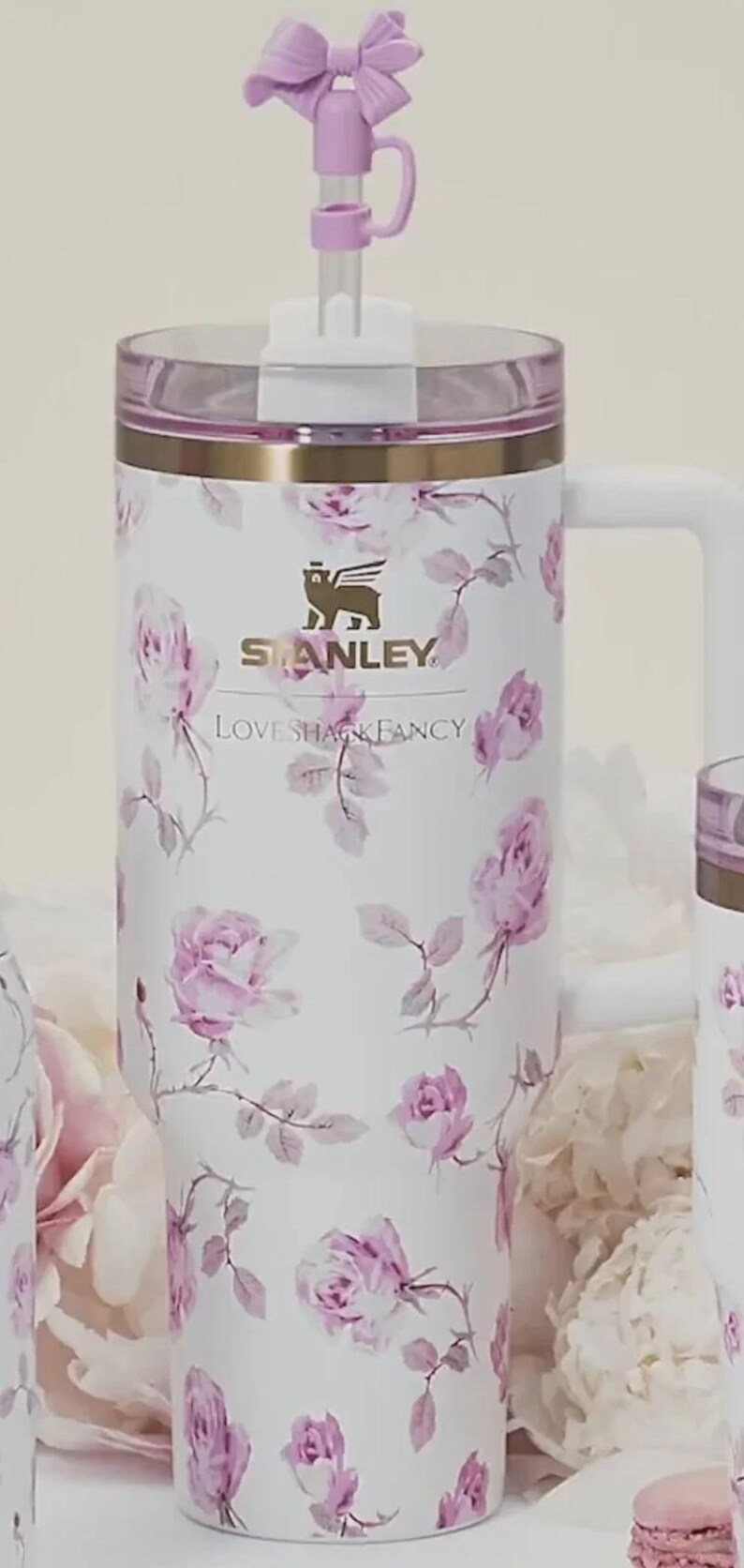Op de afbeelding: Witte Stanley-beker met een bloemenrozenpatroon in roze en grijstinten. De beker heeft een handvat, een doorzichtig deksel en een decoratieve strik op het rietje. De logo's van Stanley en Love Shack Fancy zijn zichtbaar.