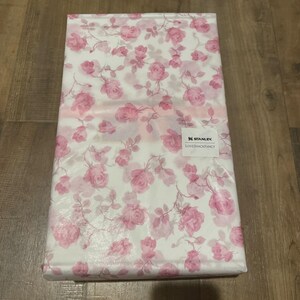 Op de afbeelding: Een gevouwen pakket verpakt in wit papier met een roze rozenpatroon. Het pakket heeft een label met de tekst "STANLEY LOVESHACKFANCY". De rozen zijn in verschillende maten en roze tinten.
