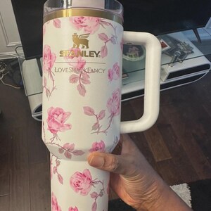 Op de afbeelding: Witte Stanley LoveShackFancy reismok met een roze rozenbloemmotief. De mok heeft een witte handgreep, een doorzichtig deksel en gouden accenten. Het Stanley-logo is zichtbaar.