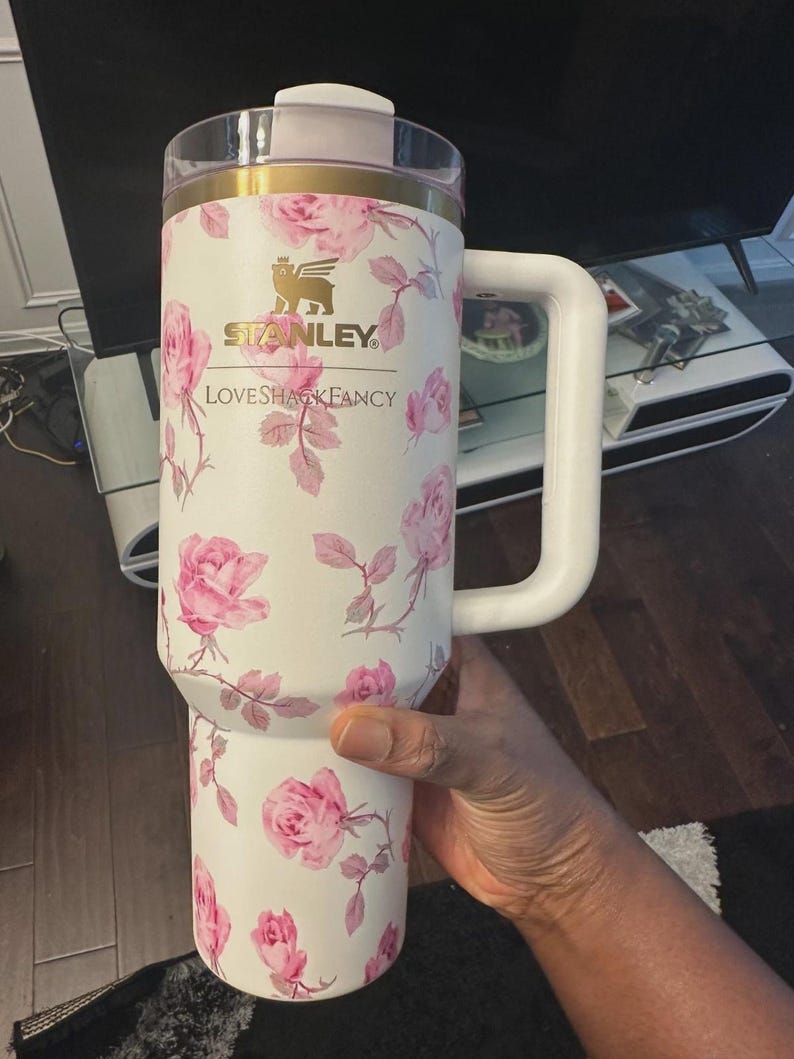 Op de afbeelding: Witte Stanley LoveShackFancy reismok met een roze rozenbloemmotief. De mok heeft een witte handgreep, een doorzichtig deksel en gouden accenten. Het Stanley-logo is zichtbaar.