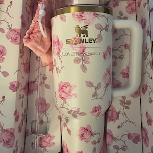 Op de afbeelding: Een witte Stanley-beker met een roze rozenpatroon, een witte handgreep en een gouden rand. De beker zit in een bloemenprint doos met een roze trekkoordtas en een doorzichtig rietje. De tekst "Stanley" en "LoveShackFancy" zijn zichtbaar.
