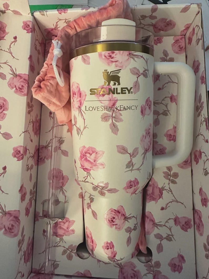 Op de afbeelding: Een witte Stanley-beker met een roze rozenpatroon, een witte handgreep en een gouden rand. De beker zit in een bloemenprint doos met een roze trekkoordtas en een doorzichtig rietje. De tekst "Stanley" en "LoveShackFancy" zijn zichtbaar.