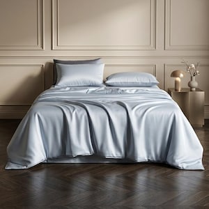 May include: A bed featuring a light blue, smooth, satin-like bedspread that drapes over the sides. Two matching light blue rectangular pillows are positioned at the head of the bed, with a dark gray pillow tucked behind them for contrast.