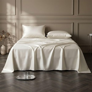 May include: A bed set featuring a smooth, cream-colored sheet draped over a mattress and two matching rectangular pillows. The fabric has a subtle sheen, and the bedding is presented in a neutral-toned room with a dark wood floor.