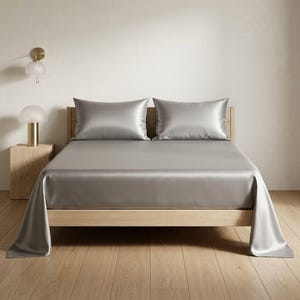Può includere: Un letto con struttura in legno chiaro è coperto da un set di lenzuola di seta grigio argento e due federe abbinate. Le lenzuola drappeggiano sui lati del letto. La stanza ha un design minimalista con pavimento in legno.