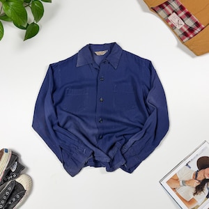 May include: A long-sleeved button-down shirt in a solid deep blue color. The shirt features a classic collar, two chest pockets, and matching blue buttons down the front placket. The fabric appears to have a soft, slightly worn texture throughout.