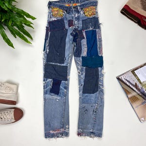 May include: Light blue denim jeans with multiple fabric patches in various shades of blue and navy. The jeans have a distressed look with paint splatters and frayed edges. The pockets are lined with a floral pattern.