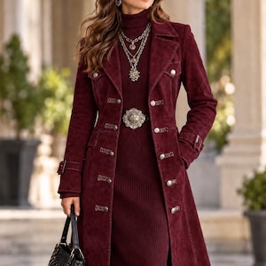 Può includere: Un trench in camoscio color bordeaux intenso con bottoni argentati, colletto strutturato e polsini con cinturino. Il cappotto è indossato sopra un abito a collo alto in maglia a coste abbinato, con una collana a catene sovrapposte e una fibbia argentata.