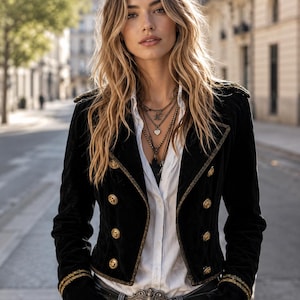 Può includere: Una giacca in velluto nero in stile militare con bottoni dorati, spalle strutturate e bordi metallici su revers e polsini. La giacca è indossata sopra una camicia bianca abbottonata, abbinata a jeans blu e una grande fibbia per cintura decorata.