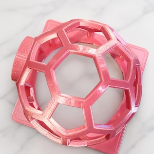 May include: A pink, geometric, spherical object with hexagonal openings, resting on a square base. The object has a soccer ball-like design and is made of a solid, possibly plastic material. The color is a vibrant, iridescent pink.
