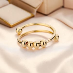 Puede incluir: Un anillo dorado con un diseño único. El anillo presenta una banda que rodea el dedo e incluye cinco pequeñas cuentas esféricas. El anillo se muestra sobre una tela suave y clara, con un joyero en el fondo.