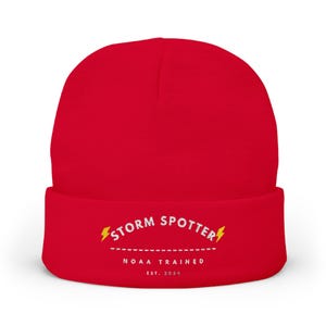 May include: A bright red cuffed beanie featuring white embroidered text that reads STORM SPOTTER with a yellow lightning bolt on each side. Below this, a dashed line sits above the text NOAA TRAINED and the smaller text EST. 2024.