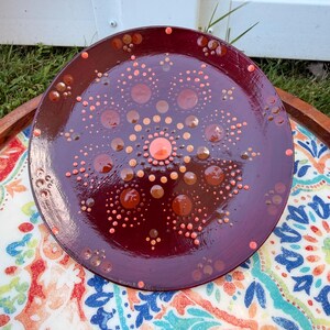 Puede incluir: Un plato redondo de color granate oscuro con un diseño de mandala pintado a mano, compuesto por numerosos puntos pequeños en relieve en tonos naranja claro, melocotón y rojo intenso. La superficie tiene un acabado brillante y el patrón irradia desde un punto central color melocotón.