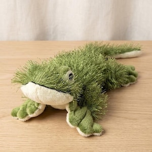 May include: A green plush frog toy with a shaggy texture. The frog has a white belly and accents, with large, round eyes. The toy is lying on a light-coloured wooden surface.