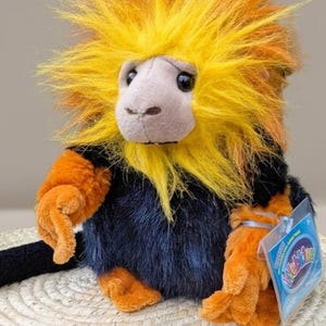 Op de afbeelding: Een knuffel van een leeuwaapje met een felgele en oranje manen, een lichtgrijs gezicht en een zwart lichaam. De aap heeft oranje ledematen en een lange zwarte staart. Aan de linkerarm van de knuffel is een blauw Webkinz-label bevestigd.