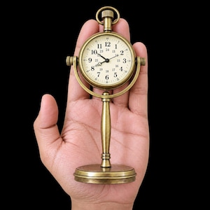 May include: A brass-toned metal desk clock featuring a round face with black Arabic numerals from 1 to 12 and smaller 24-hour markings. The clock is mounted on a vertical stand with a circular base and a top winding loop mechanism.