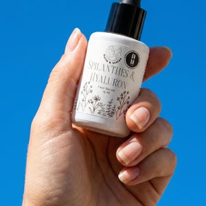 May include: A white face serum bottle with a black dropper is held against a bright blue sky. The bottle has black text that reads "SPILANTHES & HYALURON Face Serum 15 ml" with floral illustrations. The bottle is held in a hand.