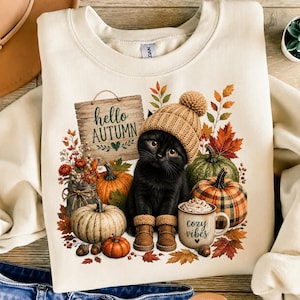 Peut inclure: Un sweat-shirt crème avec une illustration d'un chaton noir portant un bonnet et des bottes en tricot beige. Le motif comprend des citrouilles, une tasse marquée "cozy vibes" et un panneau en bois "hello AUTUMN" entouré de feuilles d'automne.