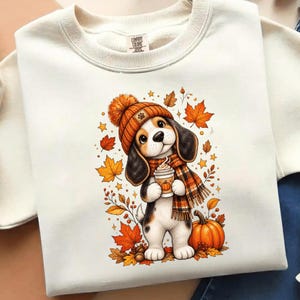 Peut inclure: Un sweat-shirt à col rond couleur crème avec une illustration d'un chiot beagle portant un bonnet en tricot orange et une écharpe à carreaux. Le chiot tient une tasse de café avec une icône de citrouille, entouré de feuilles d'automne.