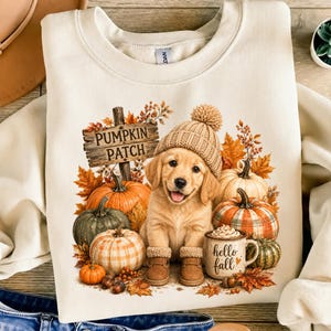 Peut inclure: Un sweat-shirt à col rond couleur crème avec une illustration d'un chiot golden retriever portant un bonnet en tricot beige. Le motif inclut des citrouilles, des feuilles d'automne, un panneau en bois PUMPKIN PATCH et une tasse hello fall.