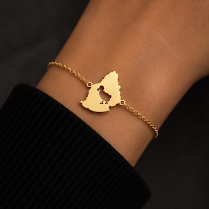 May include: A gold-toned chain bracelet featuring a central flat pendant shaped like the map of Romania. The pendant has a cutout silhouette of a small bird in the centre. The delicate metal chain connects to both sides of the map-shaped charm.