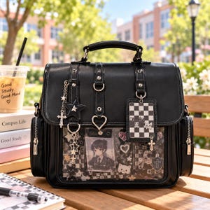 Può includere: Una borsa nera stile cartella con manico superiore e una tasca frontale trasparente che mostra illustrazioni in stile anime, hardware a forma di cuore e ciondoli a forma di stella e croce. La borsa presenta dettagli in metallo argentato.