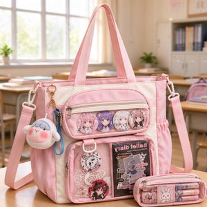 Puede incluir: Un bolso tote rosa y crema con dos asas superiores y una correa de hombro desmontable. El frente presenta un bolsillo con cremallera con cuatro pines de estilo anime, un bolsillo de malla con un diario Ballet y un dije de peluche.