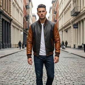 Op de afbeelding: Een varsity-jack met een zwart leren body, bruine leren mouwen, een wit t-shirt en een donkerblauwe spijkerbroek. Het jack heeft een geribbelde kraag en boorden met bruine en zwarte strepen en metalen drukknopen aan de voorzijde.