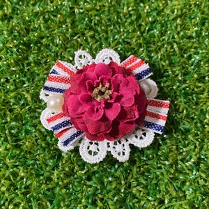 以下が含まれることがあります： 深紅色の布製フラワーモチーフに金色のアクセントをあしらった、手作りのヘアアクセサリー。白いレースのトリム、赤、白、青のストライプのリボン、2つのイミテーションパールが特徴です。緑色の背景に配置されています。