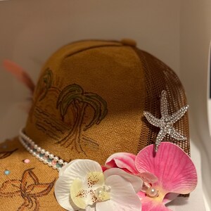 May include: A brown trucker hat with a mesh back, decorated with a rhinestone starfish, faux pearls, and vibrant pink and white orchid flowers. The hat has a painted tropical design and is embellished with small, colourful beads.