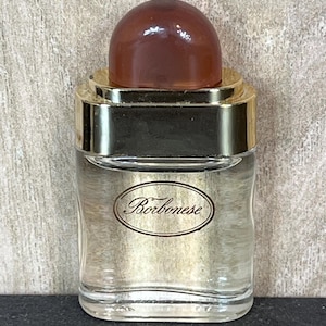 May include: A small, clear glass perfume bottle with a brown, rounded cap. The bottle has a gold-colored band around the top and the word "Borbonese" in an oval frame on the front. The bottle is filled with a clear liquid.