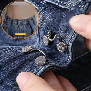 May include: A set of two silver-toned metal waist adjusters featuring a textured, grid-patterned circular design. These adjustable fasteners are shown attached to the waistband of dark blue denim jeans to tighten the fit of the garment.