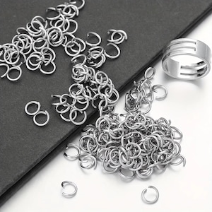 May include: A collection of small, silver-colored metal jump rings scattered across the frame, accompanied by a single, wider silver-colored metal ring featuring three horizontal cut-out lines along its outer band surface.