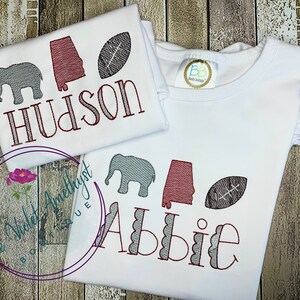May include: Two white t-shirts with embroidered designs. The first shirt has a gray elephant, a red Alabama state outline, a brown football, and the name "Hudson". The second shirt has a gray elephant, a red Alabama state outline, a brown football, and the name "Abbie".