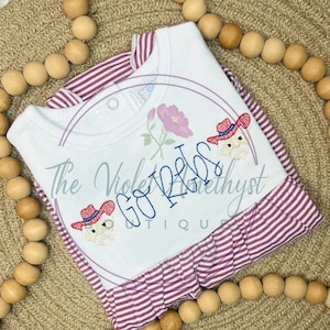 May include: White and pink striped baby dress with a white bodice featuring the text "Go Vols" in blue embroidery and two red cowboy hats with white faces embroidered on the bodice.