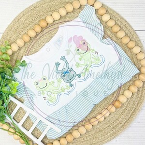 May include: A white baby dress with a blue and white striped ruffle hem. The dress features an embroidered design of three green frogs with pink bows, jumping in a pond.