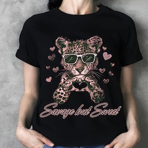 Può includere: T-shirt nera con un leopardo con occhiali da sole che forma un cuore con le zampe. Il design include cuori rosa e la scritta "Savage but Sweet" in corsivo.