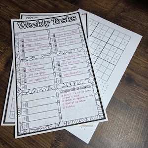 Op de afbeelding: Een stapel witte wekelijkse takenplanners en een Sudoku-puzzelblad op een houten oppervlak. De takenplanners hebben secties voor schoonmaken, eten, huishouden en een dopamine-menu. De tekst bevat "Weekly Tasks" en "Dopamine Menu".