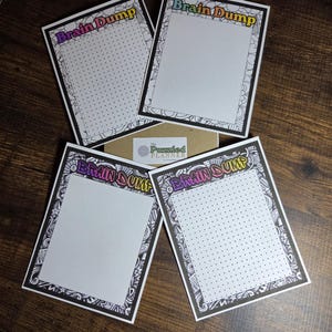 May include: Four white Brain Dump notepads with black borders and dot grids, arranged on a wooden surface. The top of each notepad has the words "Brain Dump" in colorful lettering. A small cardboard box with a logo is partially visible.