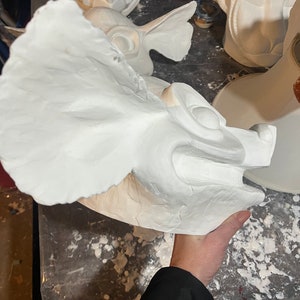 May include: A white, unfinished sculpture of a mythical creature's head with large ears and a detailed face. The sculpture is held by a person. Other similar sculptures are visible in the background.