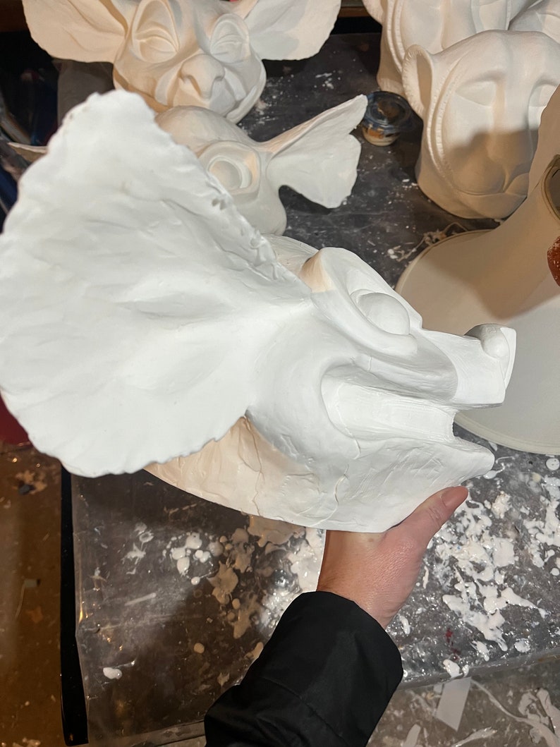 May include: A white, unfinished sculpture of a mythical creature's head with large ears and a detailed face. The sculpture is held by a person. Other similar sculptures are visible in the background.