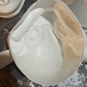 May include: A close-up view of a white, mask-like sculpture with a smooth, glossy interior. The sculpture is partially filled with a white, liquid substance, possibly paint or resin. The background is a dark surface with paint splatters.
