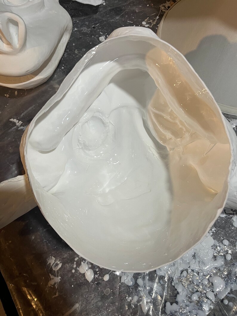May include: A close-up view of a white, mask-like sculpture with a smooth, glossy interior. The sculpture is partially filled with a white, liquid substance, possibly paint or resin. The background is a dark surface with paint splatters.