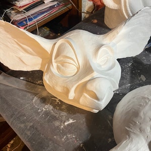 Op de afbeelding: Een close-up van een witte, onafgewerkte sculptuur van een dierenkop met grote oren en gesloten ogen. Andere soortgelijke sculpturen zijn gedeeltelijk zichtbaar op de achtergrond. De stijl suggereert een grillig of fantasiethema.