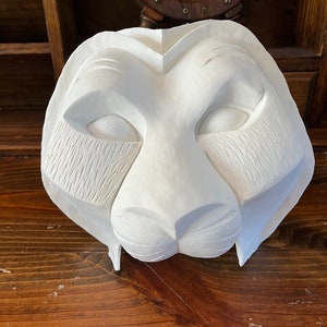 Puede incluir: Máscara de león blanca tallada con detalles faciales, incluyendo ojos cerrados y detalles de pelaje texturizado. La máscara está hecha de un material sólido y se muestra sobre una superficie de madera. Objeto decorativo.