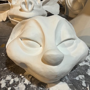 May include: A collection of white, unfinished plaster masks. The masks feature various animal-like faces with closed eyes, prominent noses, and rounded cheeks. Some masks have large ears. The masks are on a dark surface with white paint splatters.